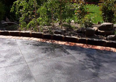 Curved patio with dark gray tiles and red brick edging, next to a garden with plants and rocks.