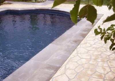 A swimming pool with dark blue water, surrounded by light beige tiles and a stone-textured deck, with greenery in the foreground.