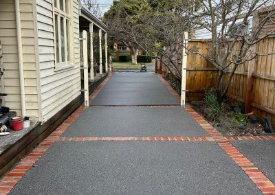 A neat concrete pathway bordered by brick accents, flanked by a house on one side and a wooden fence with trees on the other.