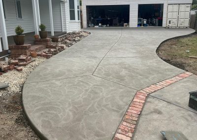 A newly paved concrete driveway curves toward a garage, featuring a brick path and landscaped edges with potted plants and rocks.
