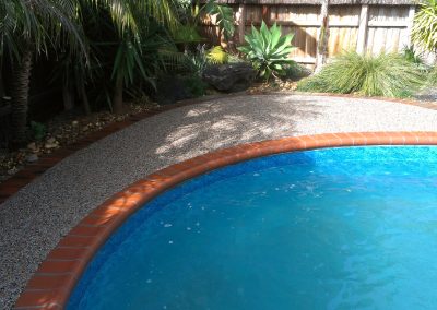 A clear blue swimming pool surrounded by a textured stone deck, greenery, and a wooden fence in a sunny backyard setting.