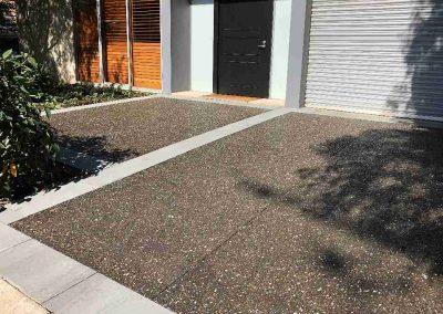A modern entrance features a sleek gray door and polished gravel driveway, surrounded by greenery and wooden slatted panels.