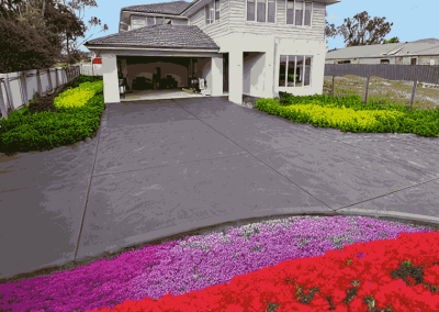 A modern house with a gray driveway, bordered by colorful flower beds featuring red, pink, and green plants.