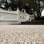 The charm of exposed aggregate and picket fences Close-up of a pebble-textured driveway leading to a white picket fence with trees in the background.