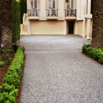 The depth and style of exposed aggregate makes it the pre-eminent concrete choice A pebble-textured driveway leading to a cream-colored building with balconies, bordered by palm trees and greenery.