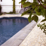 This sandstone design and light colour work perfectly A rectangular swimming pool next to a patterned stone walkway and a white building.