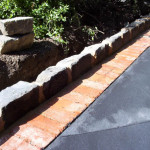 We can create bluestone and brick border for a beautiful garden edge A driveway with a stone and brick border next to a plant bed with bushes.