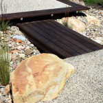 Around rocks or against timber, exposed aggregate is unequalled. Modern landscape with wooden footbridge, pebble path, and large stone.