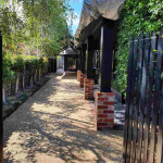A landscaped pathway with bushes on one side and a fence on the other, leading to a house with a black facade and a metal roof.
